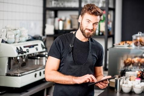 Ein junger Barista mit Bart steht in einem Caf&eacute; und bedient ein Tablet, umgeben von Kaffeezubeh&ouml;r und Geb&auml;ck.