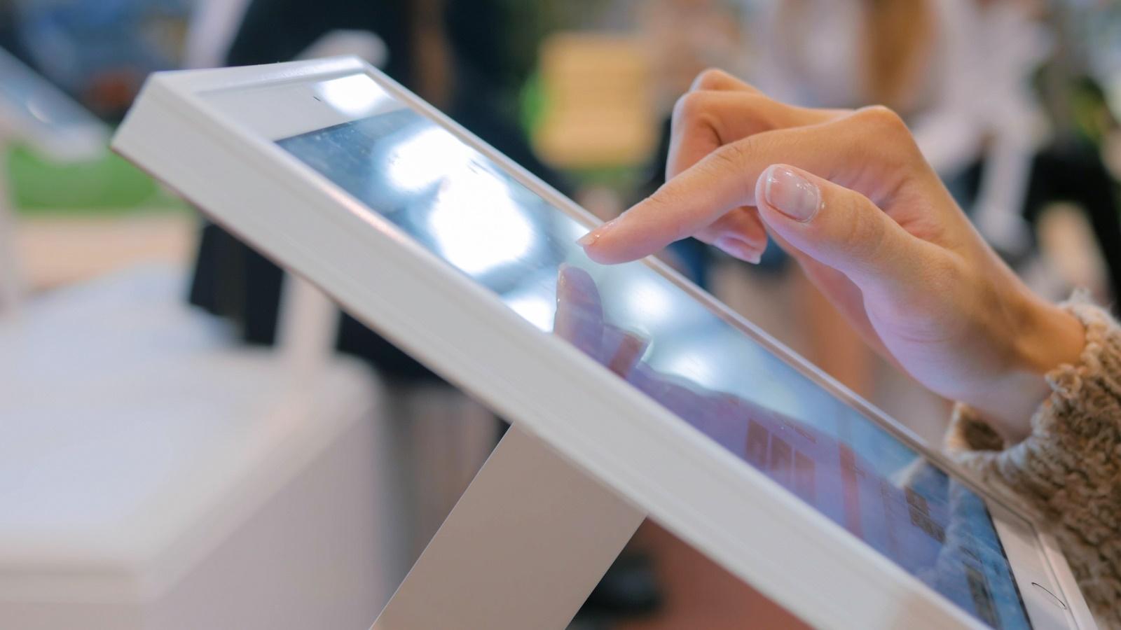 Eine Hand tippt auf einen modernen, wei&szlig;en Touchscreen-Monitor in einem hellen, &ouml;ffentlichen Raum.