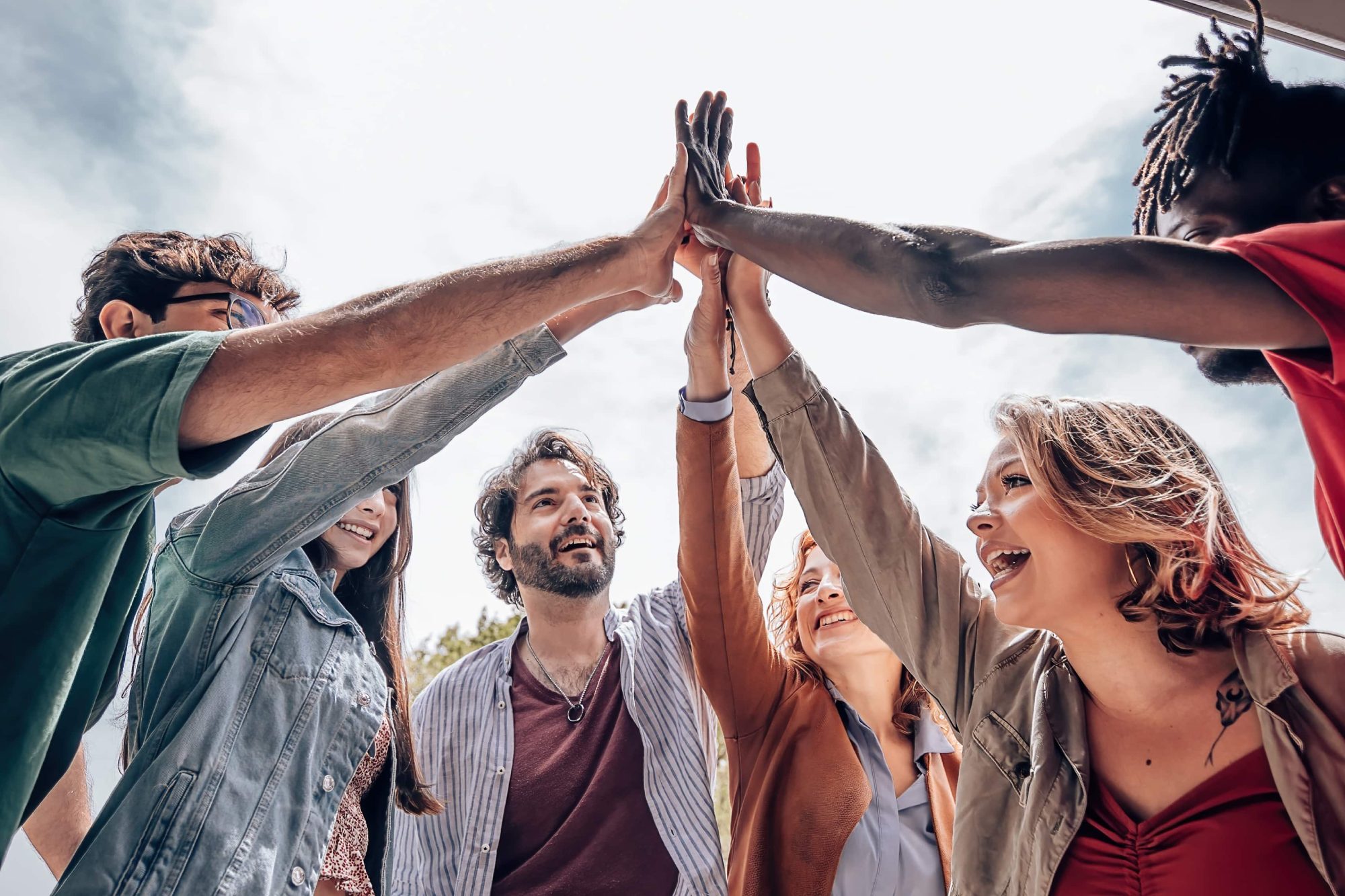 Sechs Menschen feiern den Erfolg, indem sie ihre Hände in die Mitte heben, unter einem hellen Himmel.