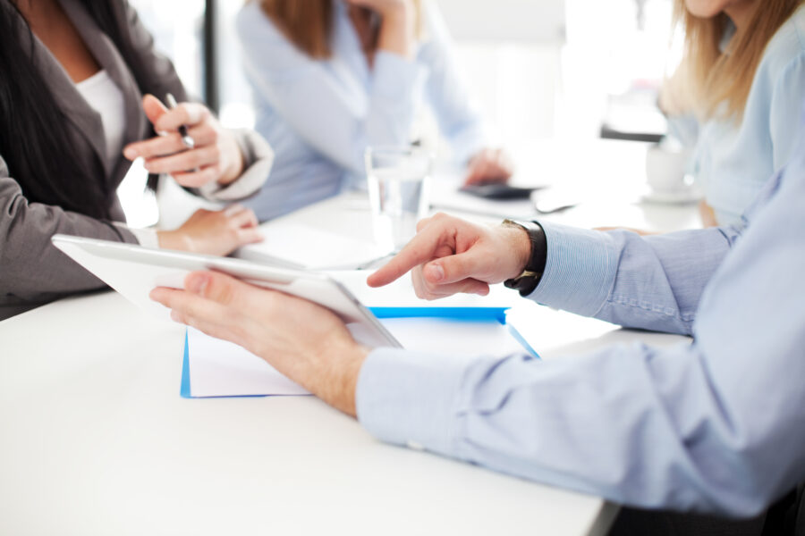 Eine Person zeigt auf ein Tablet w&auml;hrend einer Besprechung am Tisch mit weiteren Teilnehmern.