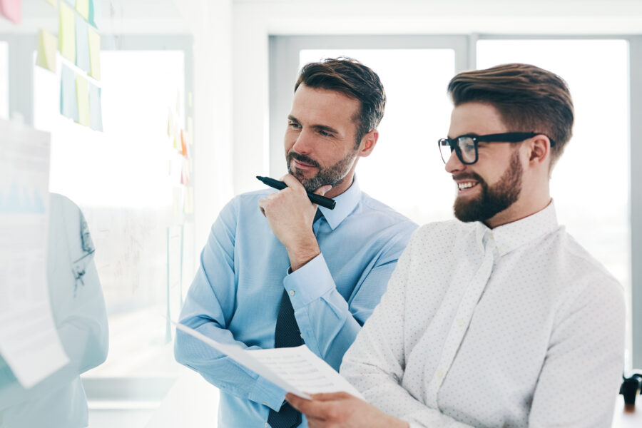 Zwei M&auml;nner in B&uuml;rokleidung, einer nachdenklich mit Stift, der andere l&auml;chelnd, betrachten Notizen an einer Wand.