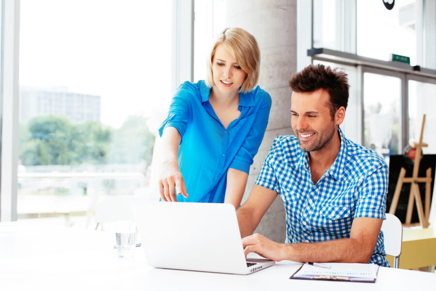 Ein Mann und eine Frau arbeiten an einem Laptop, w&auml;hrend sie sich in einem modernen B&uuml;ro unterhalten.