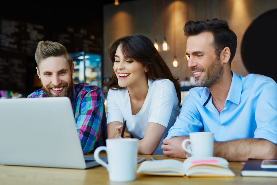 Drei Personen sitzen an einem Tisch in einem Caf&eacute; und schauen gemeinsam auf einen Laptop. Tassen stehen vor ihnen.