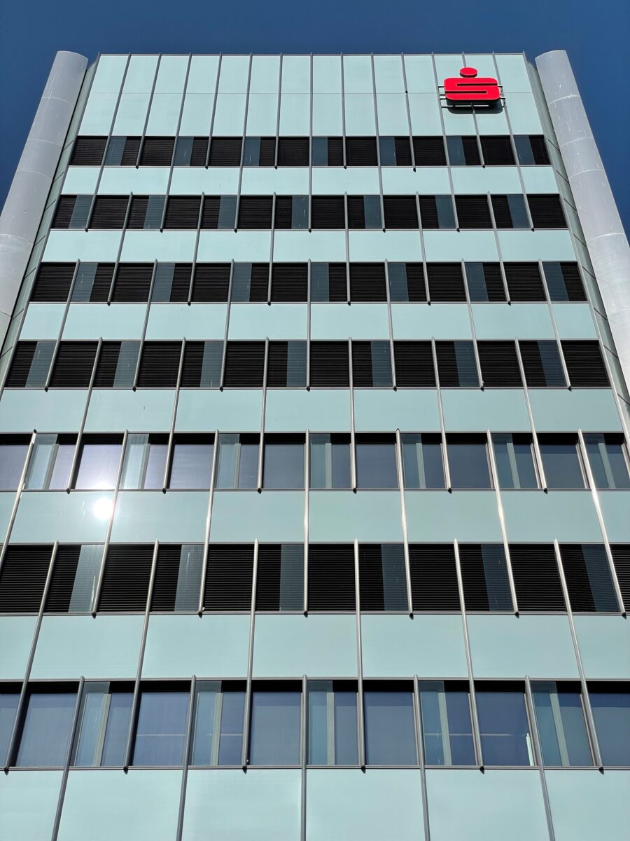 Modernes Geb&auml;ude mit reflektierenden Fenstern und einem roten Logo oben. Klare blaue Himmel im Hintergrund.