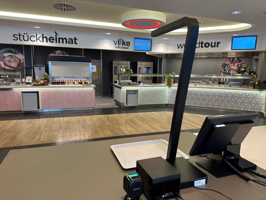 Moderne Kantine mit verschiedenen Essensstationen und digitaler Anzeige, im Vordergrund ein Kassensystem.