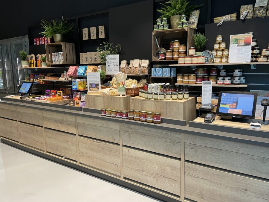 Ein Verkaufsstand mit Regalen voller Lebensmittelprodukte, Gl&auml;sern, Snacks und einer Kasse in modernem Design.