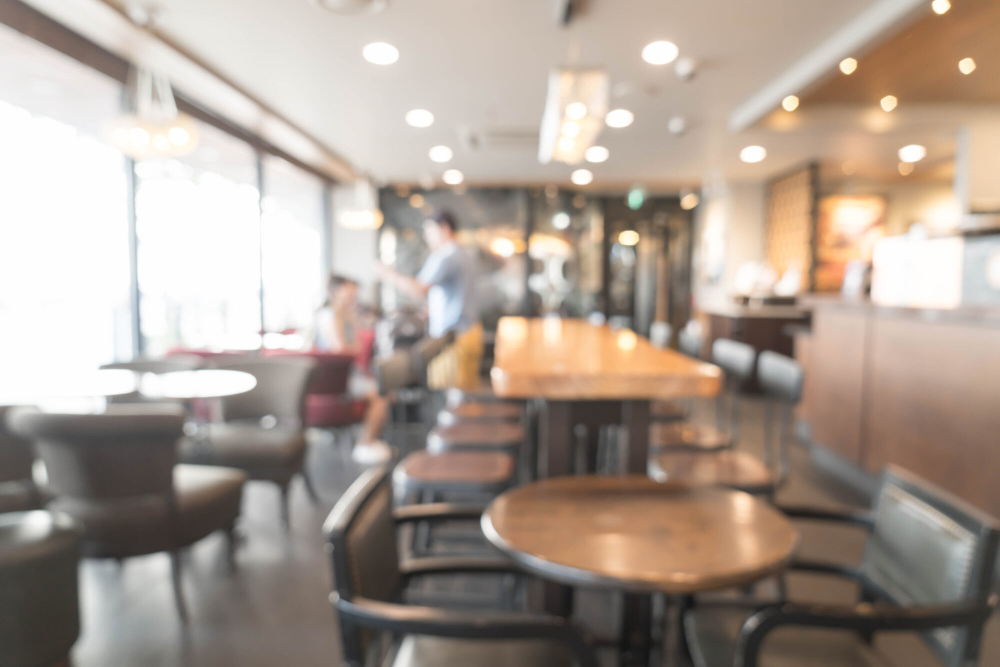 Unscharfe Darstellung eines Cafés mit Tischen, Stühlen und Menschen im Hintergrund. Helle, einladende Atmosphäre.